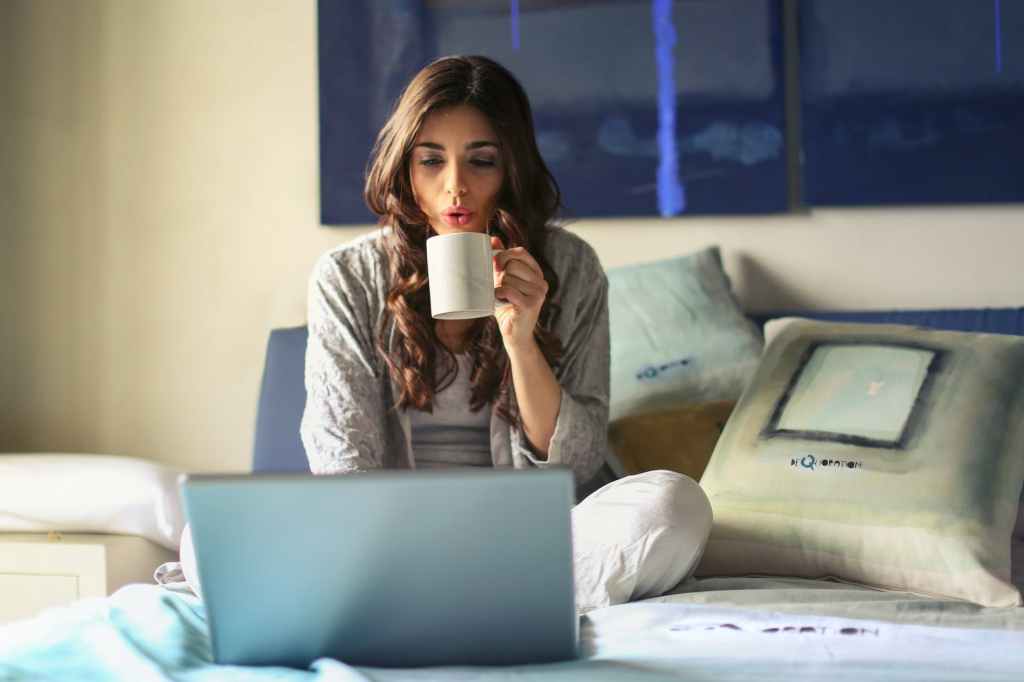 woman in grey jacket sits on bed uses grey laptop